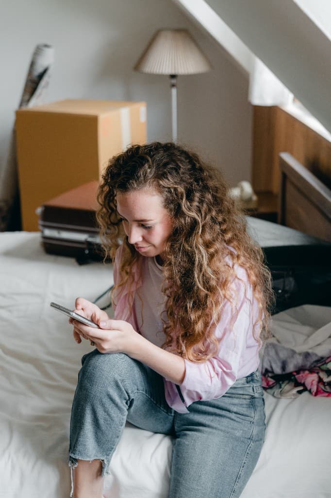 Femme consultant son téléphone sur un lit, entre cartons et valise
